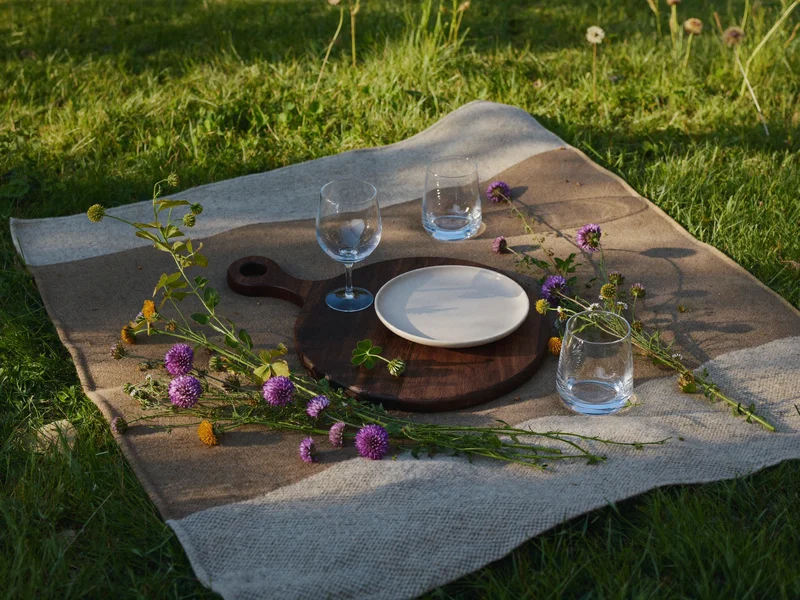 Plaid déplié sur l'herbe avec table dressée, planche en bois, verres et fleurs